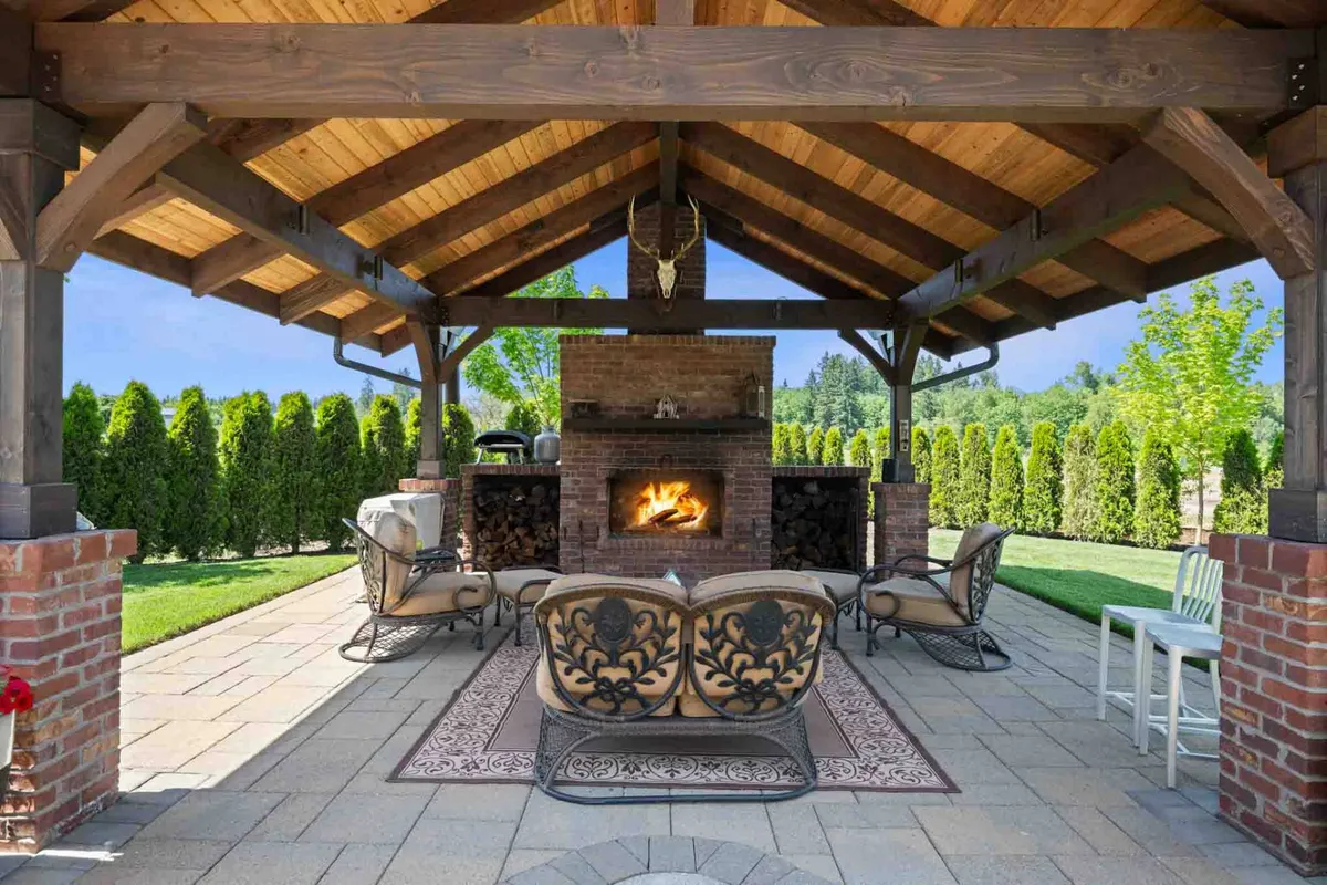 The exterior scene features a cozy pergola-covered fireplace and extensive seating area surrounded by greenery. There's an outdoor kitchen with a BBQ, perfect for al fresco dining, with sunset lighting that adds warmth and invites social gatherings.