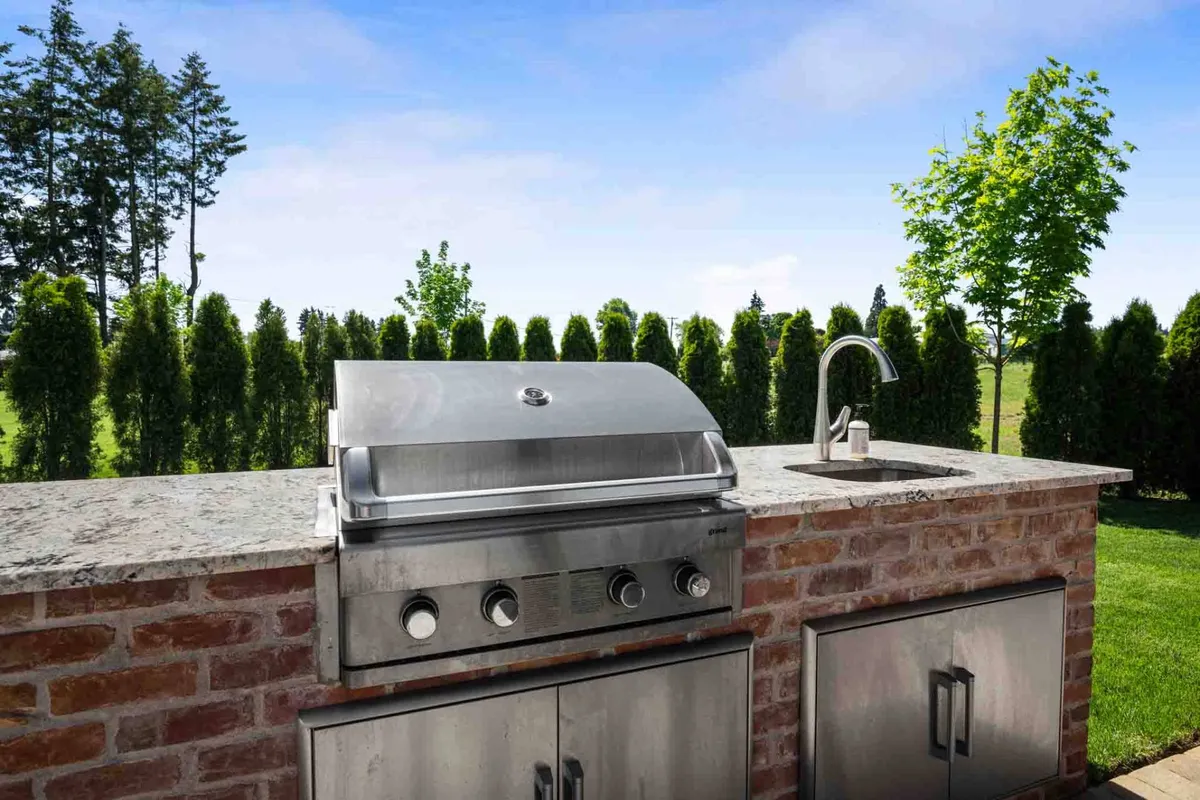 This exterior scene spotlights a stainless steel grill set against a stone countertop and lush landscaped gardens, forming an inviting space for outdoor cooking and barbecues with friends and family. The vibrant, social mood fosters both relaxation and festivity.