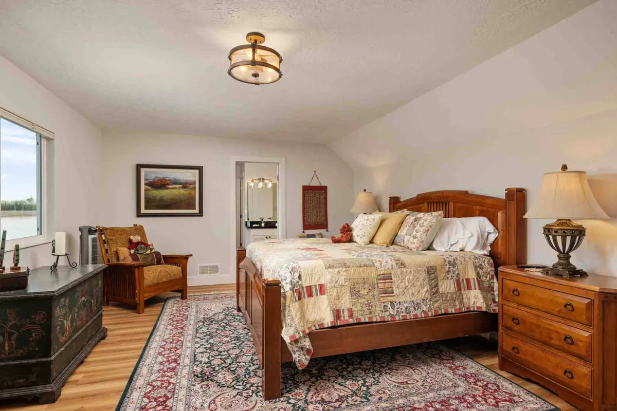 This image features a serene bedroom with a plush bed, cozy rug, sunlit window, elegant artwork, and a nightstand. The atmosphere is calm and welcoming, ideal for rest and relaxation.
