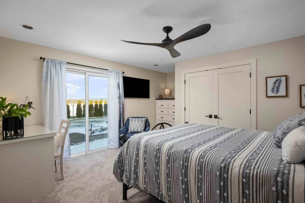 A serene bedroom with striped bedding, a sliding glass door that brings in natural light, and a cozy work nook with a desk and chair. The atmosphere is restful and flexible, ideal for relaxation or productivity.