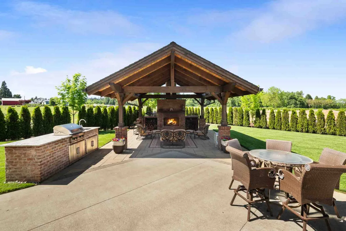 This image features a spacious wooden pergola with a built-in outdoor fireplace, a large dining table set up for gatherings, a dedicated grill and food prep area, and manicured hedges providing privacy. The scene is set for sunset dining, socializing by the fire, and enjoying the outdoors in upscale comfort.