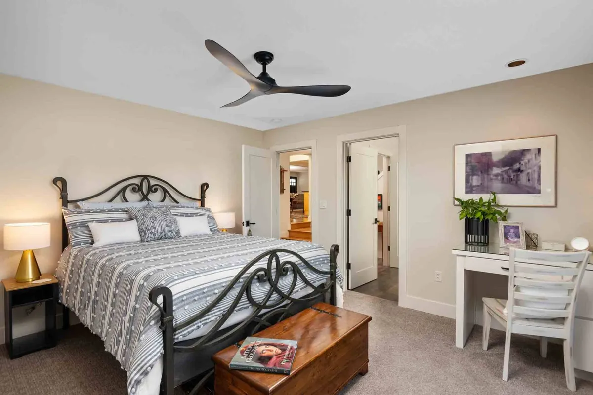 A beautifully appointed bedroom with decorative linens, a modern ceiling fan, wooden trunk at the bed’s foot, and a desk with chair, offering both relaxation and a workspace for personal time.