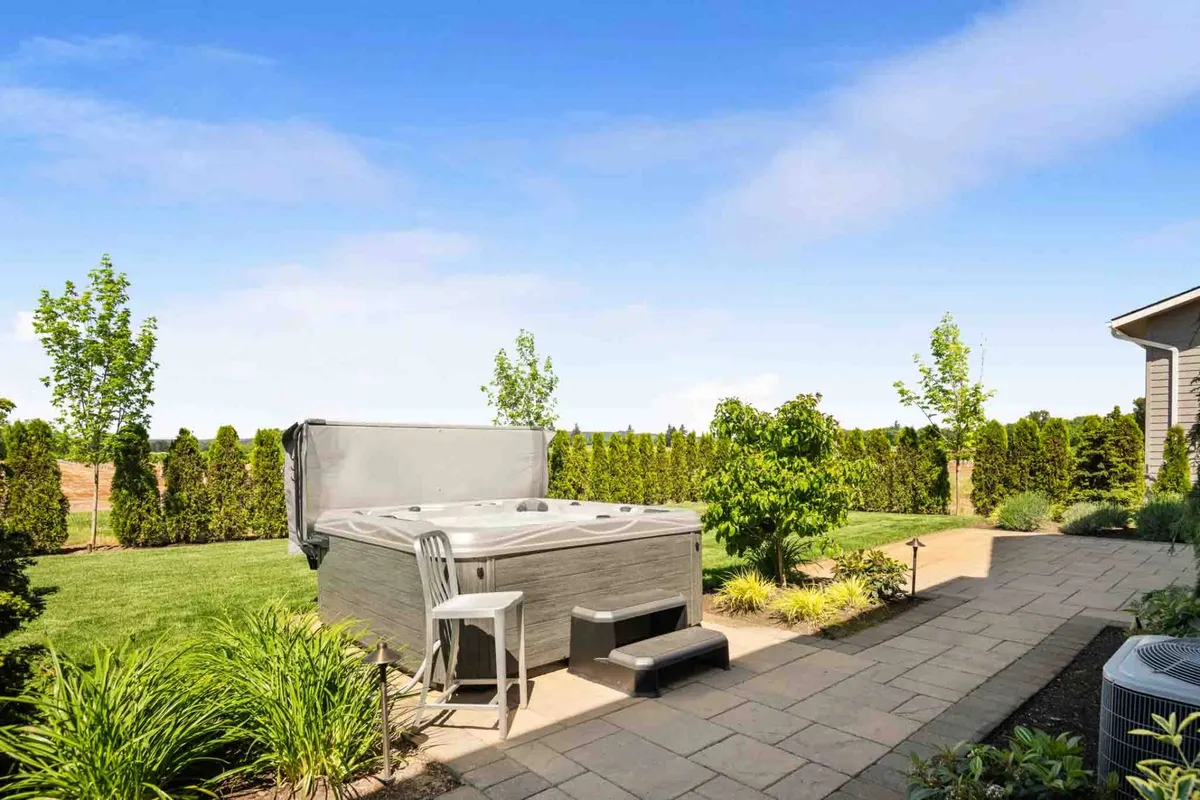 A tranquil patio scene with a hot tub, outdoor furniture, lush greenery, and ample space for entertaining. The warm lighting and spacious layout invite guests to unwind, entertain, and enjoy the outdoors.